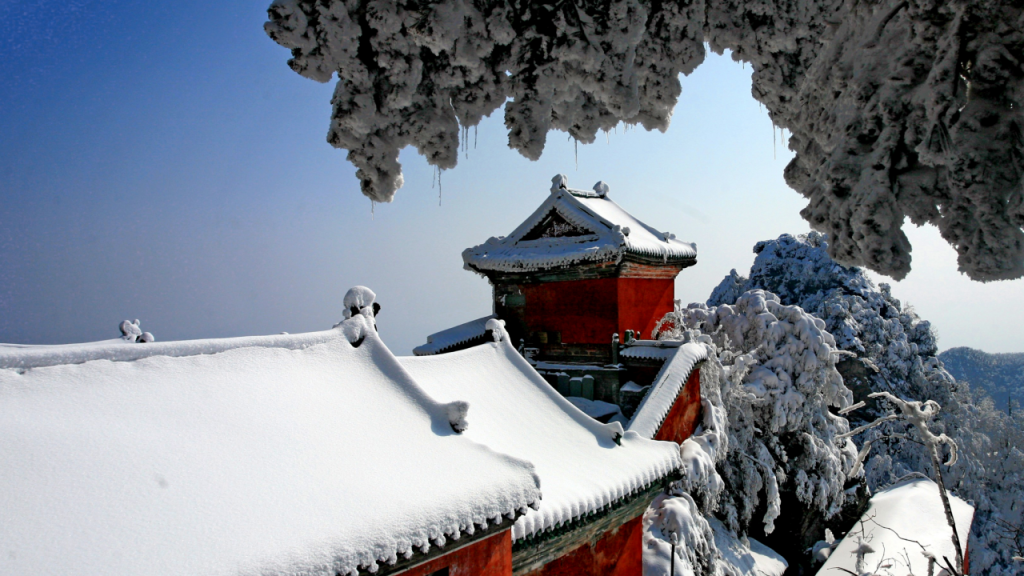 The traditional swooping roofs and deep red walls of the Taihe Palace complex covered in heavy winter snow, framed by frozen, ice-capped trees.